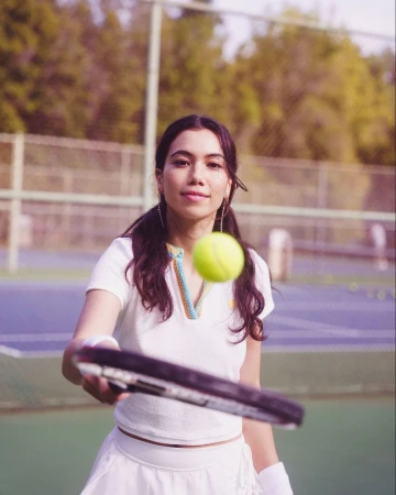 Woman in tennis whites on a blue tennis court holding a racquet with a tennis ball in the air, captured in medium shot in daylight.