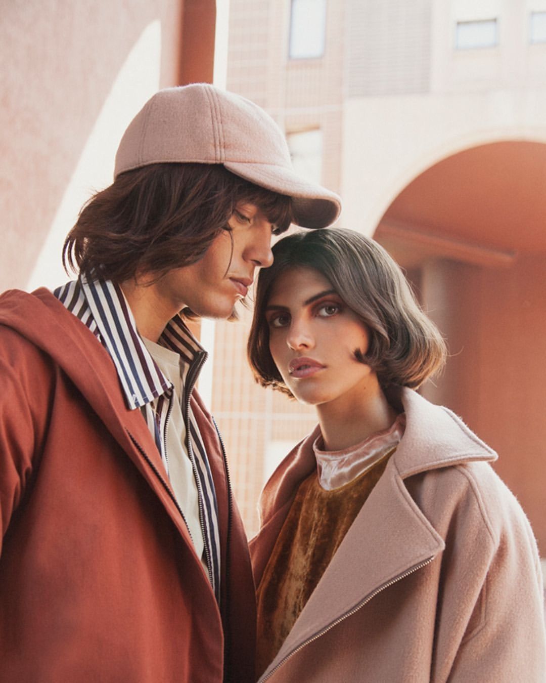 Two fashion models in a posed portrait: a man in a baseball cap and terracotta jacket, and a woman in a pink overcoat.