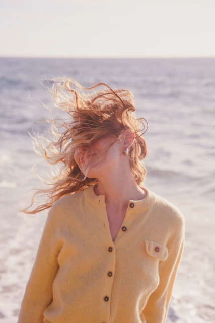 Young woman with windswept strawberry blonde hair stands on a beach in front of ocean, wearing a yellow cardigan sweater.