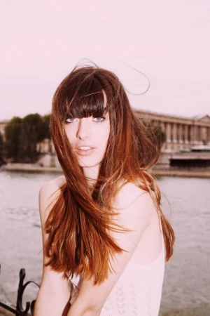 Outdoor portrait of a young adult female with long auburn hair, smiling slightly, posing in front of a canal and a Parisian building.