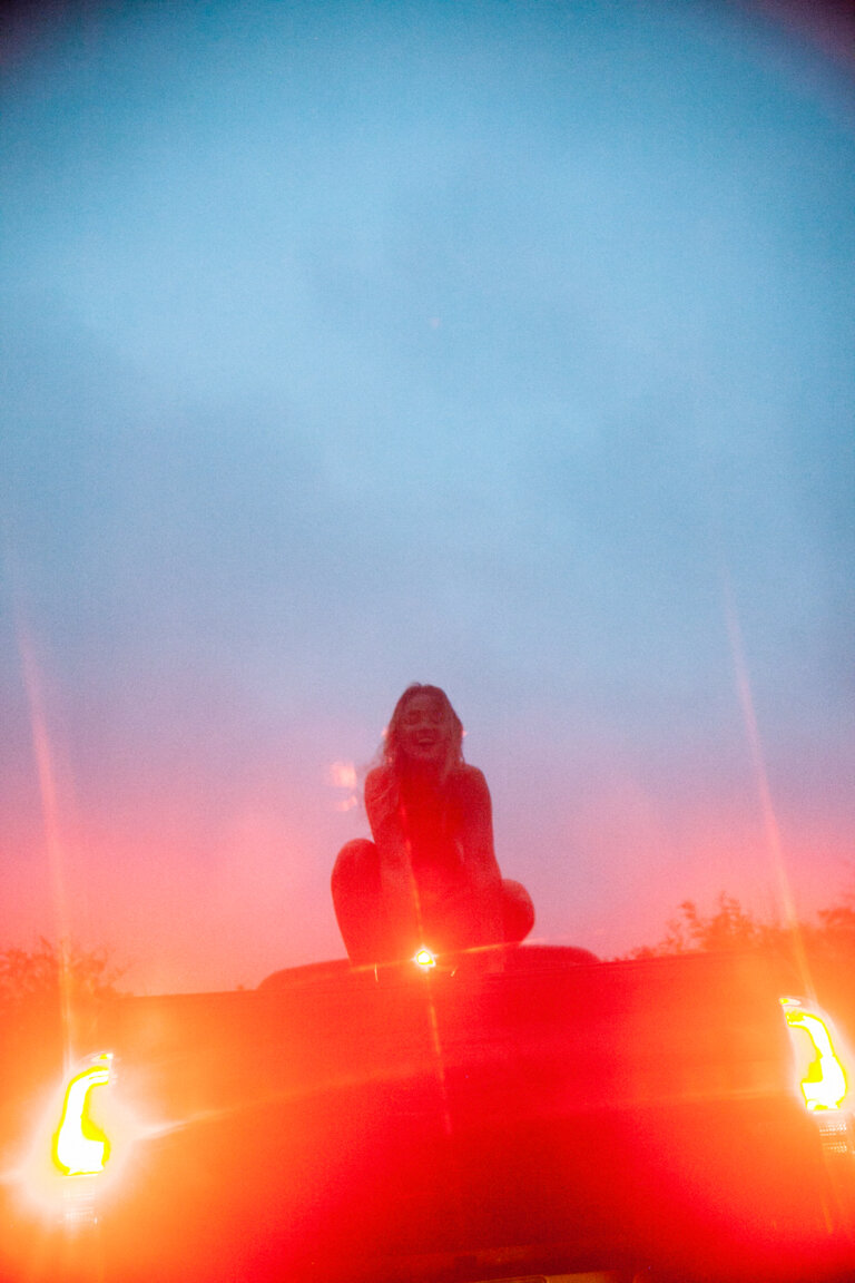 A young woman sits on the hood of a red car at dusk, smiling with hands near her face. The sky is a mix of blue and orange.
