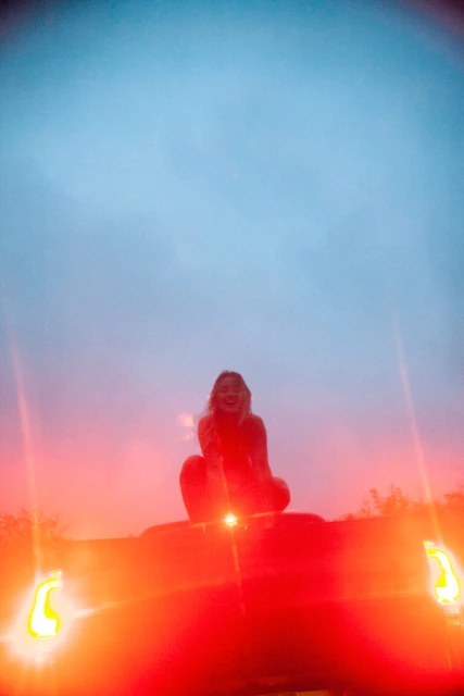 A young woman sits on the hood of a red car at dusk, smiling with hands near her face. The sky is a mix of blue and orange.