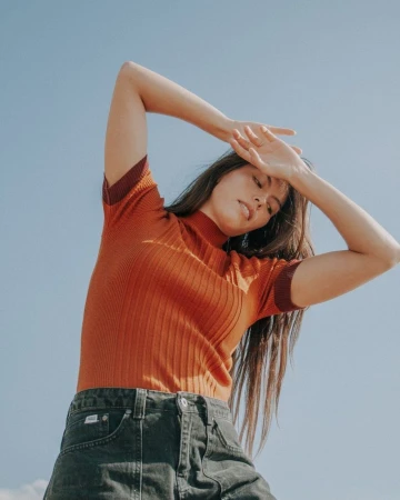 Woman in ribbed rust-orange short-sleeve top and dark denim jeans posing against clear blue sky by Nastasia Dusapin