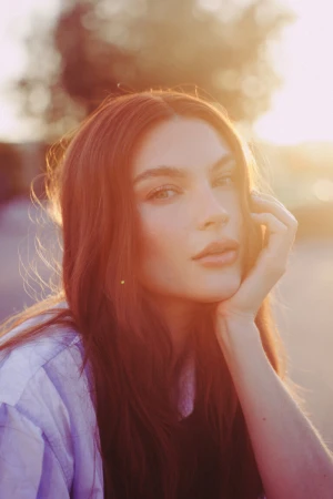 Close-up portrait of a woman with long dark hair resting her hand on her cheek outdoors in bright golden hour sunlight.