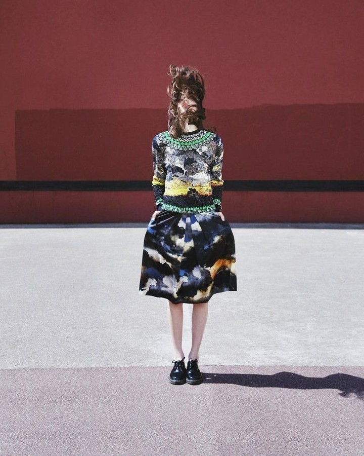 A woman stands in a watercolor patterned skirt and matching top in front of a red wall, her face obscured by her windblown brown hair.