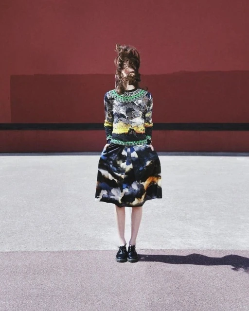 A woman stands in a watercolor patterned skirt and matching top in front of a red wall, her face obscured by her windblown brown hair.