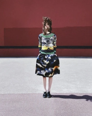 A woman stands in a watercolor patterned skirt and matching top in front of a red wall, her face obscured by her windblown brown hair.