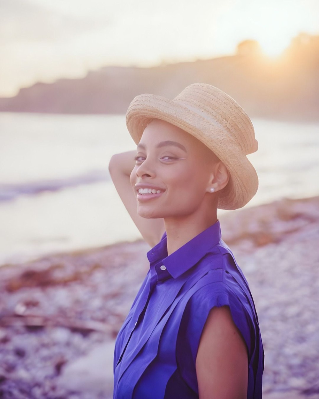 Portrait of a smiling adult female on a pebble beach in a blue top and straw hat, backlit by golden sunlight during the golden hour.