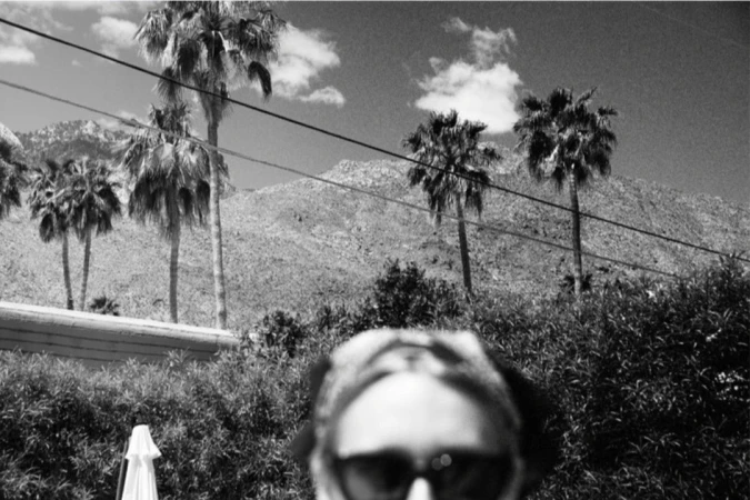 Monochrome film photograph showing the top of a person's head and sunglasses against palm trees and mountains