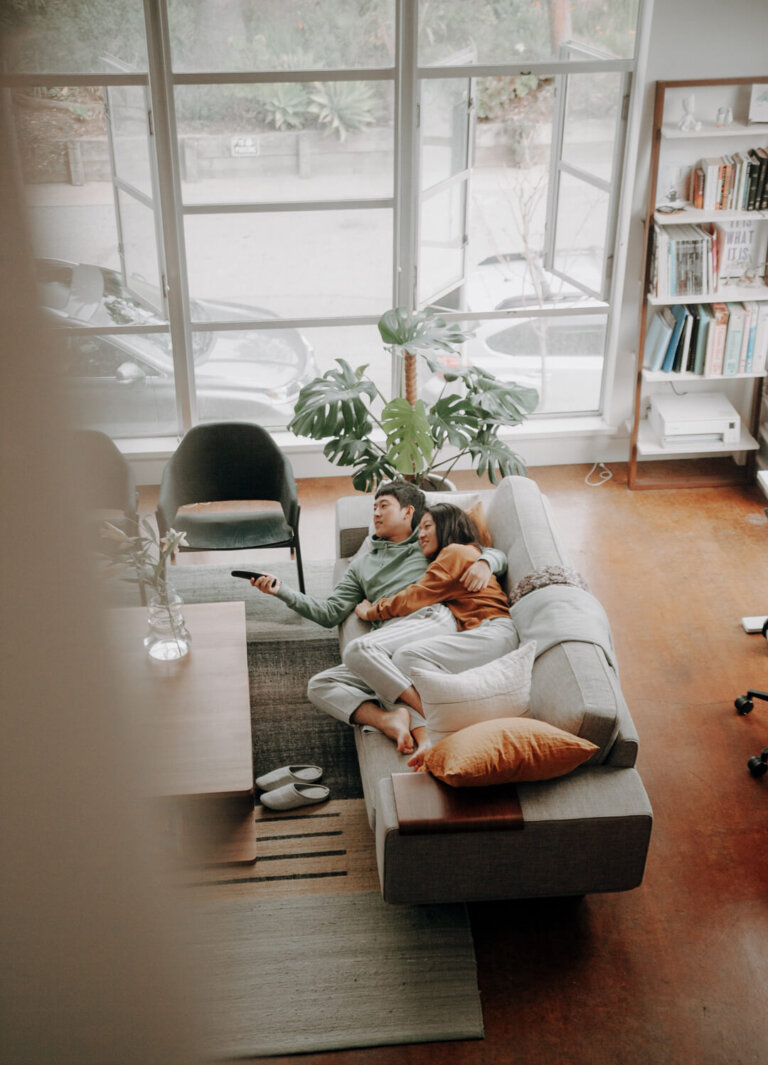 Couple relaxes on a sofa in a modern living room with large windows, indoor plants, and soft, natural light during the daytime.