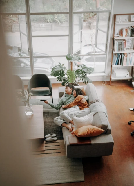 Couple relaxes on a sofa in a modern living room with large windows, indoor plants, and soft, natural light during the daytime.