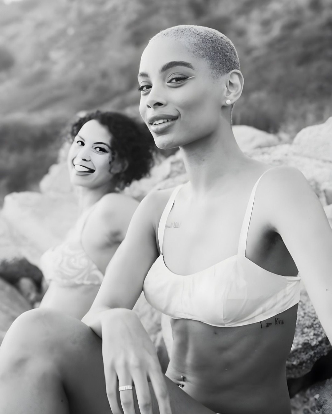 Two women pose on a rocky beach in a black and white shot. One woman has short hair and a bra top, with a second woman in the background.