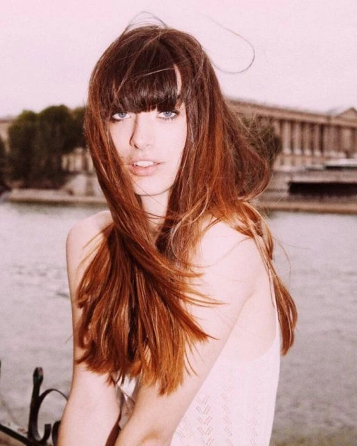 Close-up of a young woman with auburn hair and bangs, fair skin, and blue eyes, outdoors in an urban setting near a river.