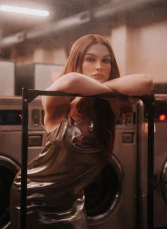 Woman with long auburn hair wearing a shiny slip dress leaning on a clothing rack inside a laundromat.