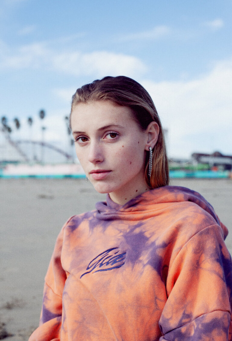 A young woman with fair skin poses on a beach wearing a tie-dye hoodie, with a water and sky background and palm trees in the distance.