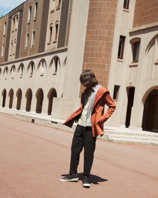 A person stands in front of a light-colored building with archways, wearing a striped shirt, orange jacket, and black pants, posing outdoors.