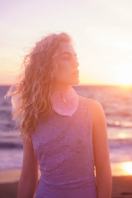 Young woman with curly hair wearing a lacy sleeveless dress and a pale pink Anthurium flower stands on a beach at sunset.