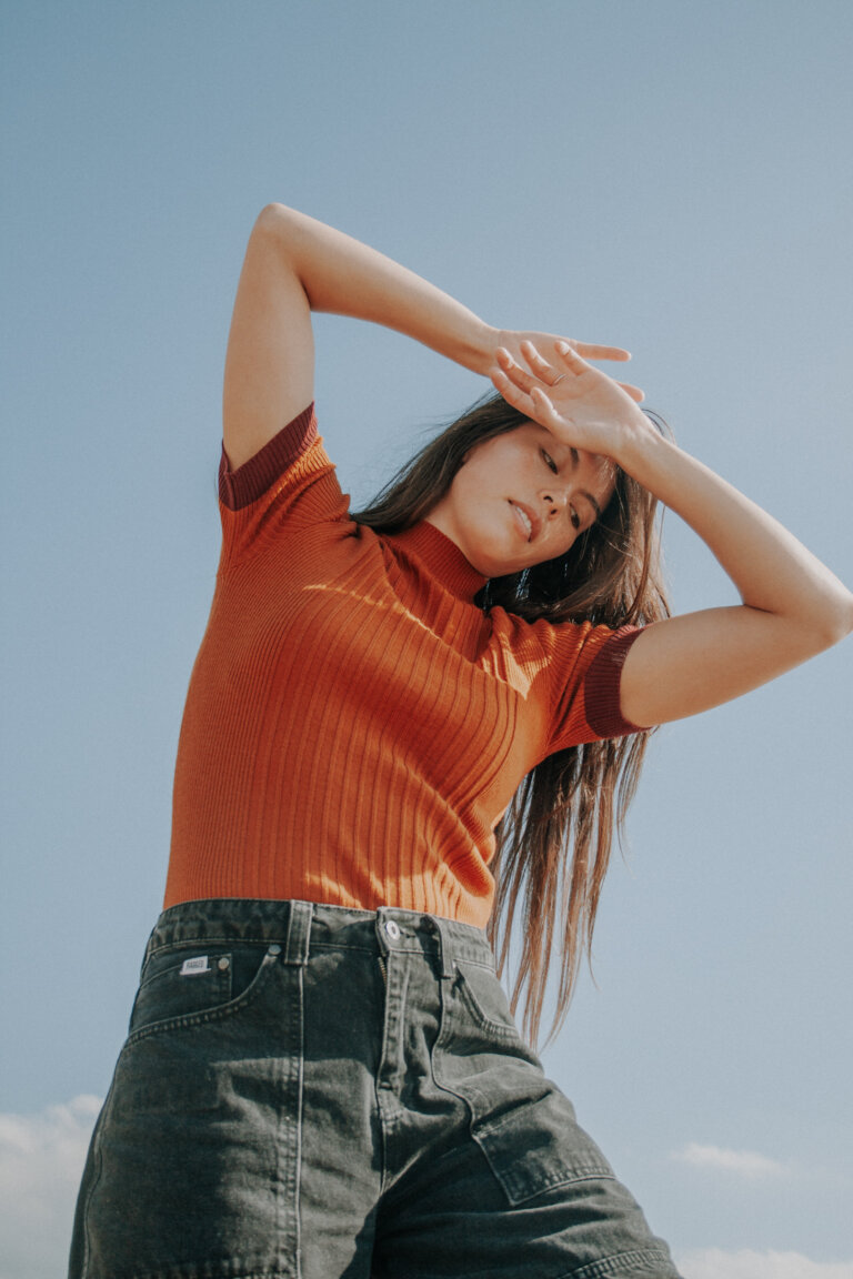 Woman in orange top and dark pants posing in front of blue sky, arms above head, eyes closed, outdoor natural light fashion shot.