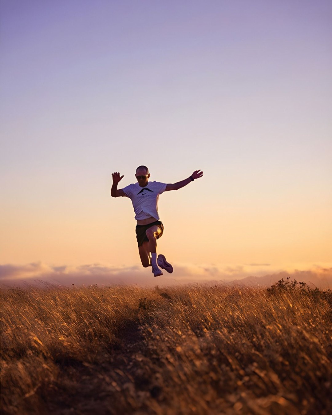 An adult male leaps across a golden field at sunset, wearing sunglasses, a white t-shirt, and dark shorts with an orange sky in the background.