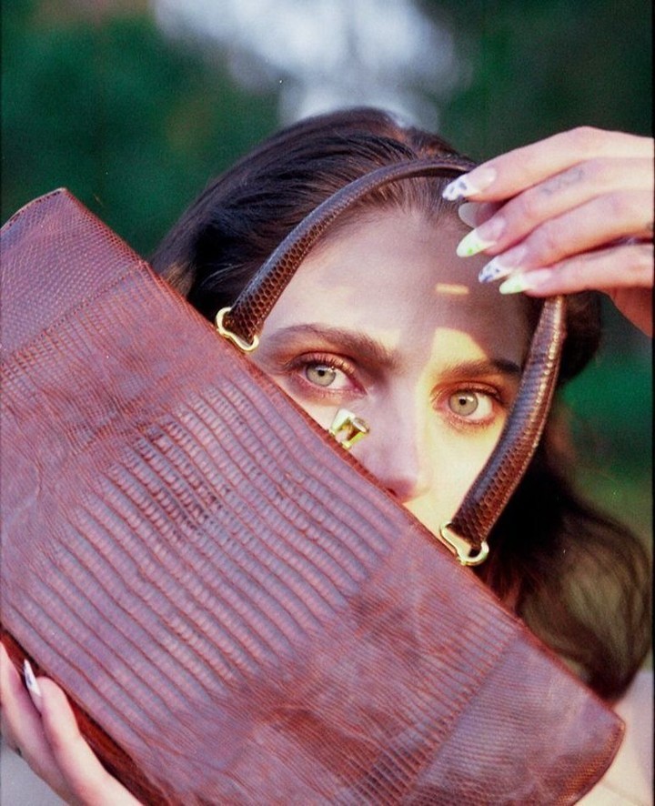 Portrait of a female peering through a brown purse with reptile texture, nails painted, with a blurred green foliage background.
