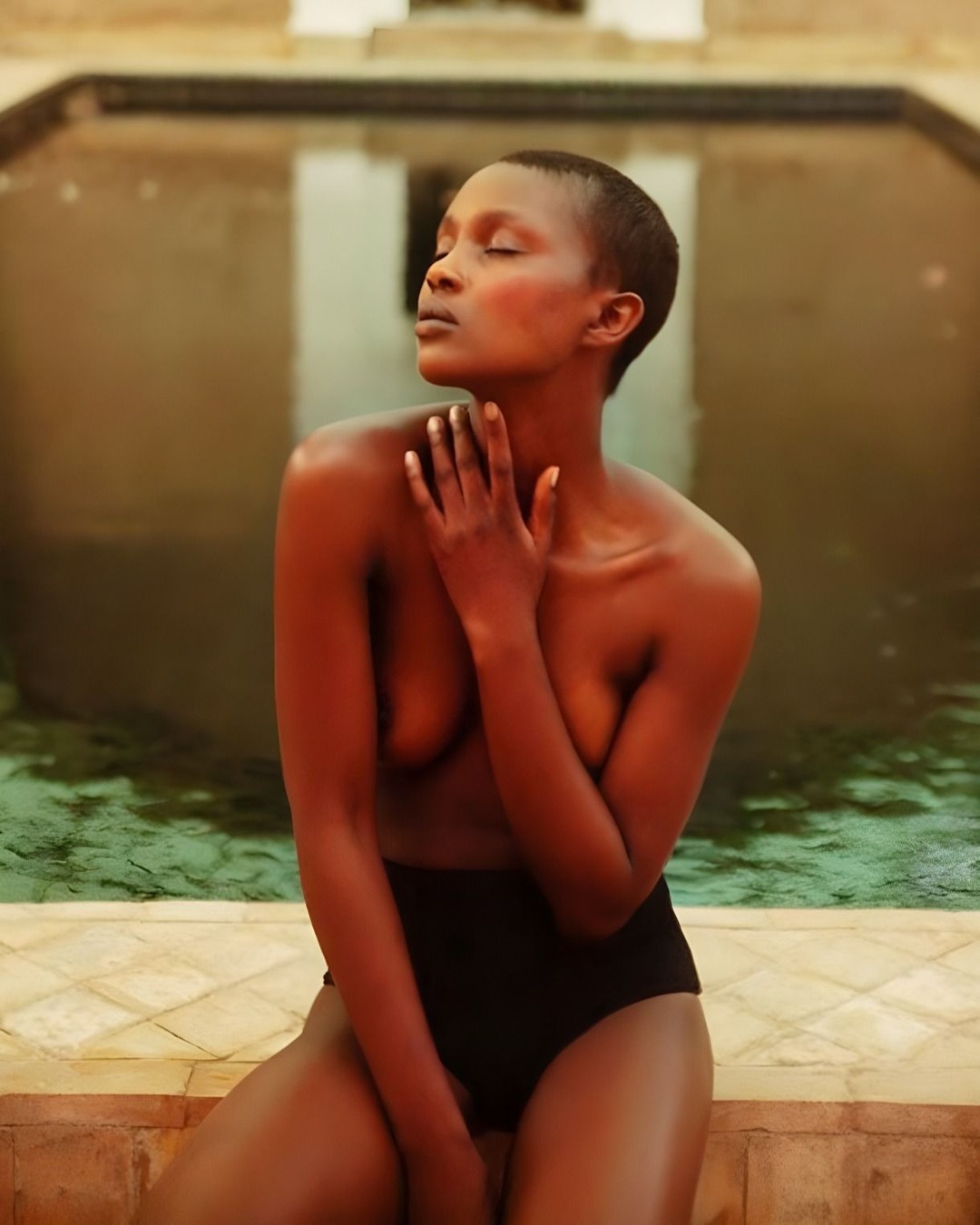 A woman with dark hair, wearing black bottoms, poses against a blurred background of a pool and stone architecture in the sunlight.