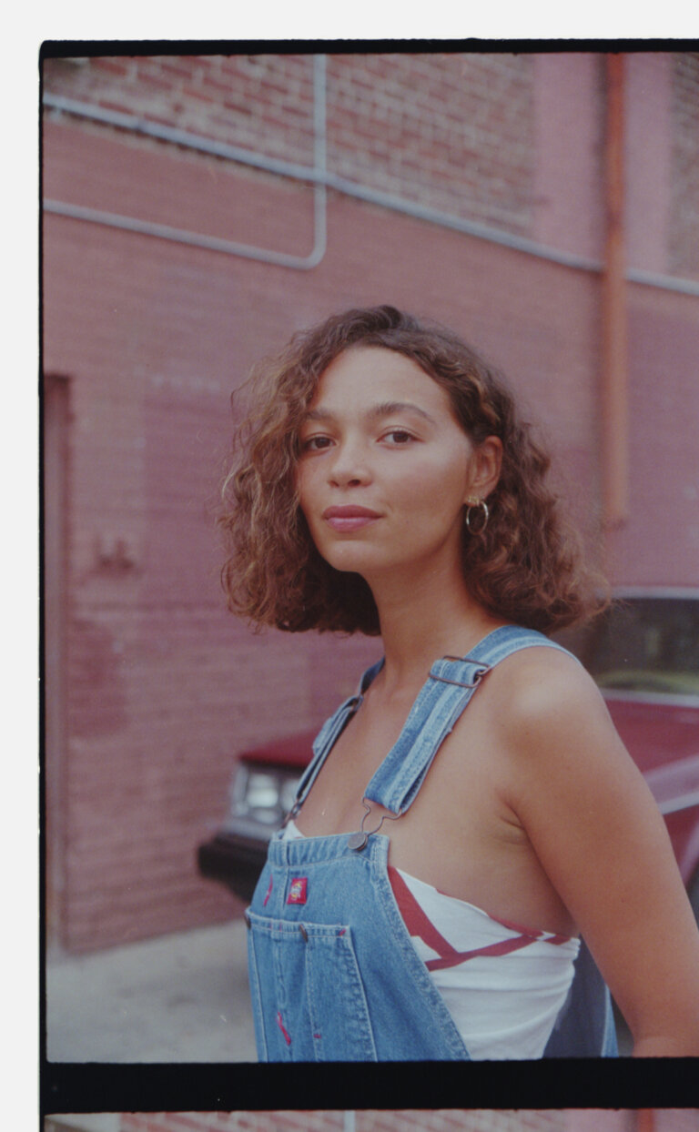 Portrait of a woman wearing denim overalls standing against a brick wall with a vintage car in the background by Nastasia Dusapin