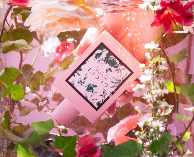 Gucci Bloom fragrance bottle held in hand among flowers, leaves, and water droplets against a pink gradient background.