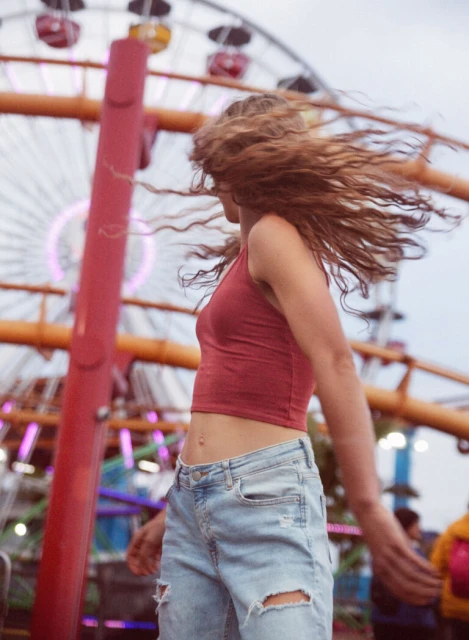 A young woman with long wavy hair, wearing a red top and jeans, dances in front of a colorful ferris wheel at an amusement park.