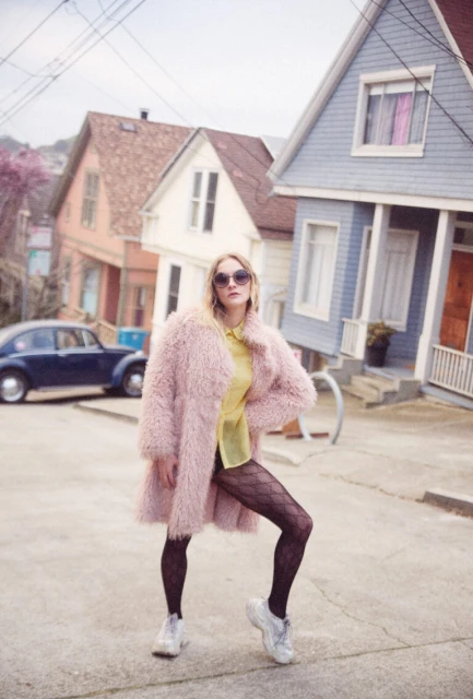 A blonde woman poses in a pink coat, yellow blouse, and black tights on a sloping street with pastel houses and a classic car.