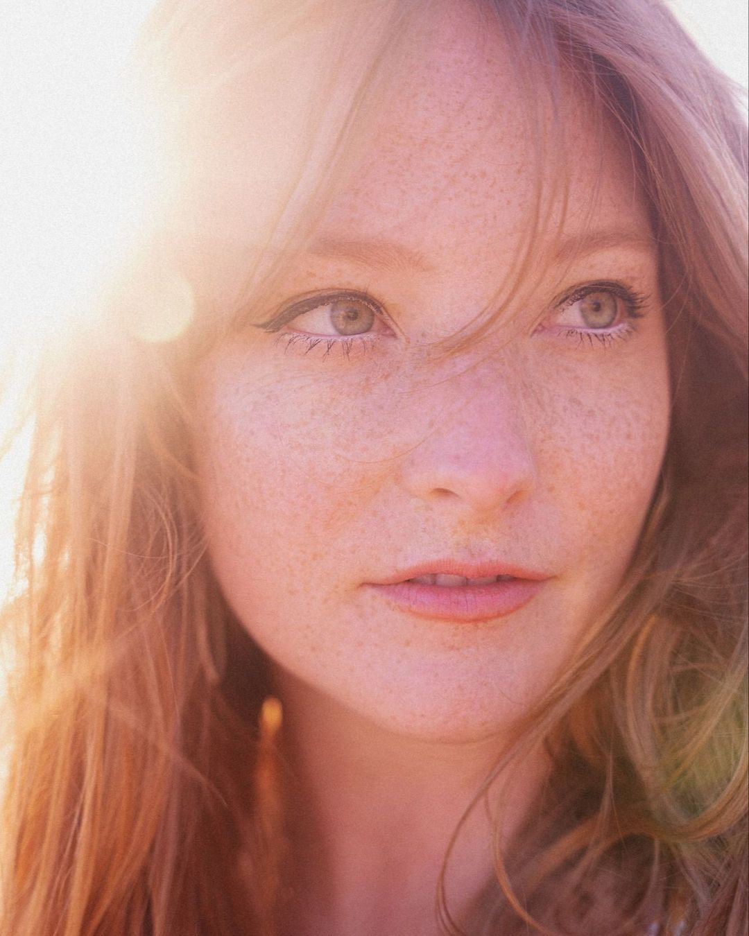 Close-up portrait of a young adult woman with freckles, fair skin, and red hair, looking off-camera to the right with soft lighting and a gentle expression.