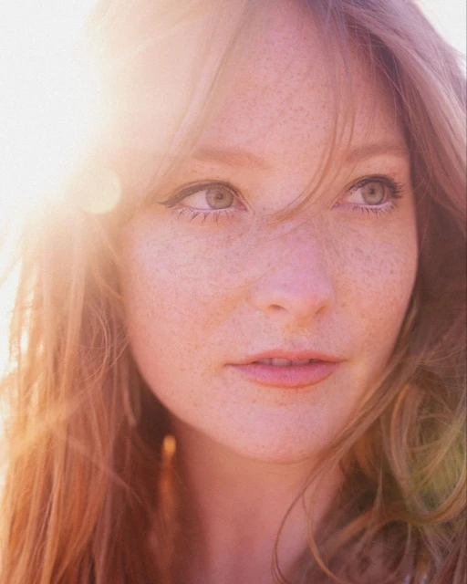 Close-up portrait of a young adult woman with freckles, fair skin, and red hair, looking off-camera to the right with soft lighting and a gentle expression.