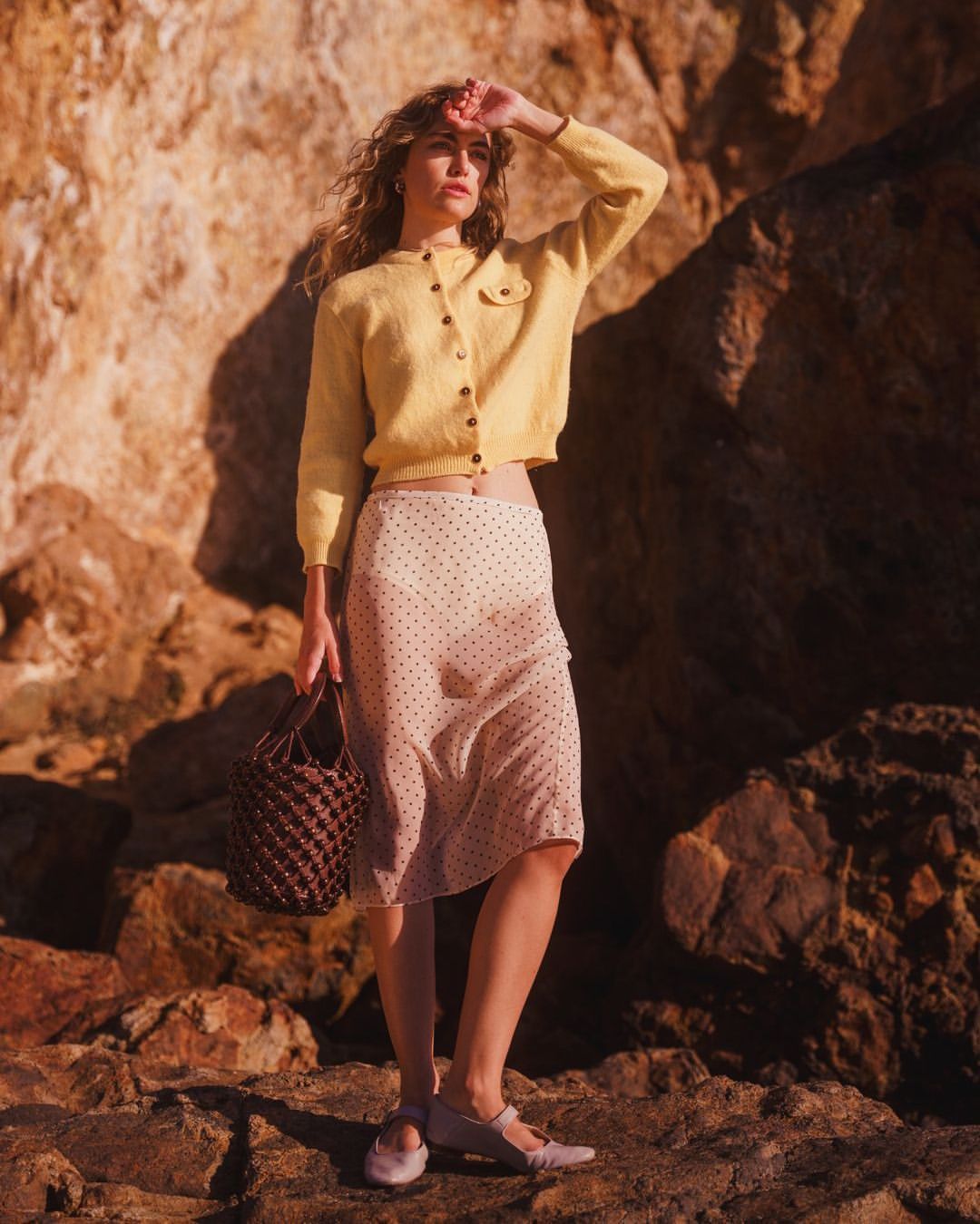 Woman in yellow cardigan and polka dot skirt stands on rocks holding a woven bag and shielding her eyes from the sunlight.