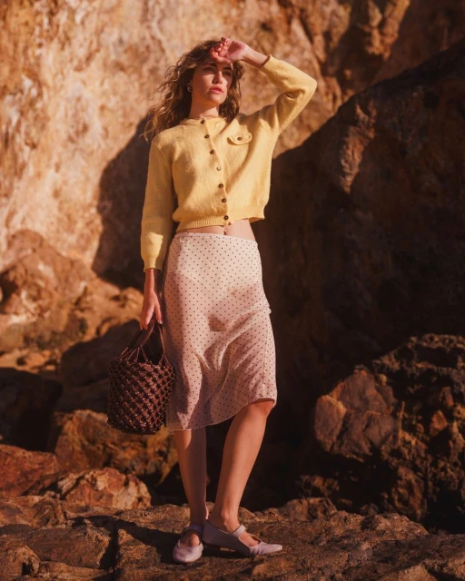Woman in yellow cardigan and polka dot skirt stands on rocks holding a woven bag and shielding her eyes from the sunlight.