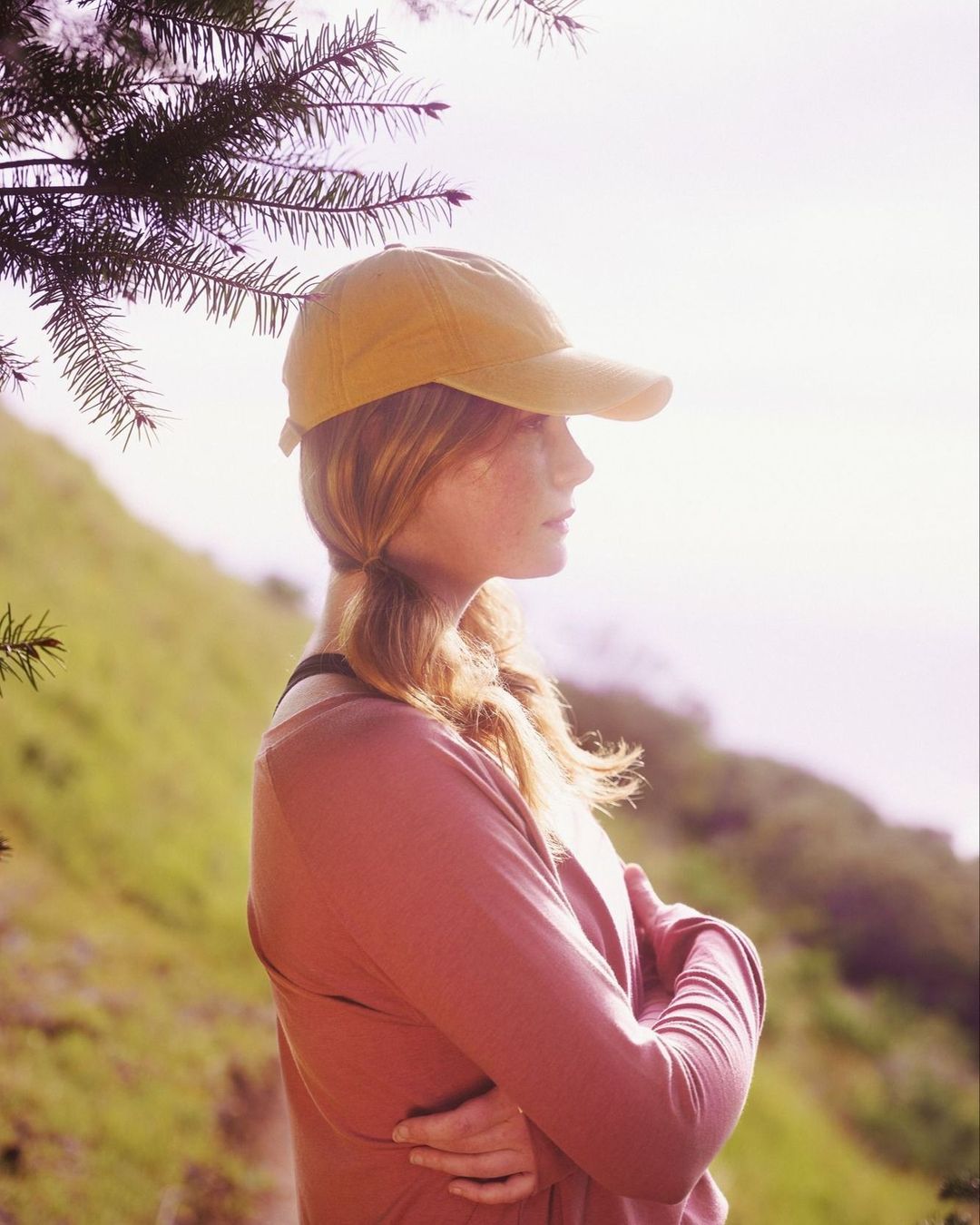 Profile of a woman with red hair wearing a cap, standing outdoors with crossed arms against a green hillside backdrop.