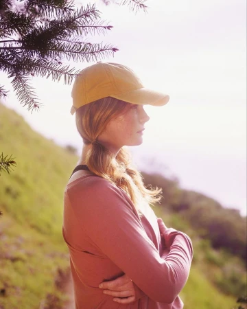 Profile of a woman with red hair wearing a cap, standing outdoors with crossed arms against a green hillside backdrop.