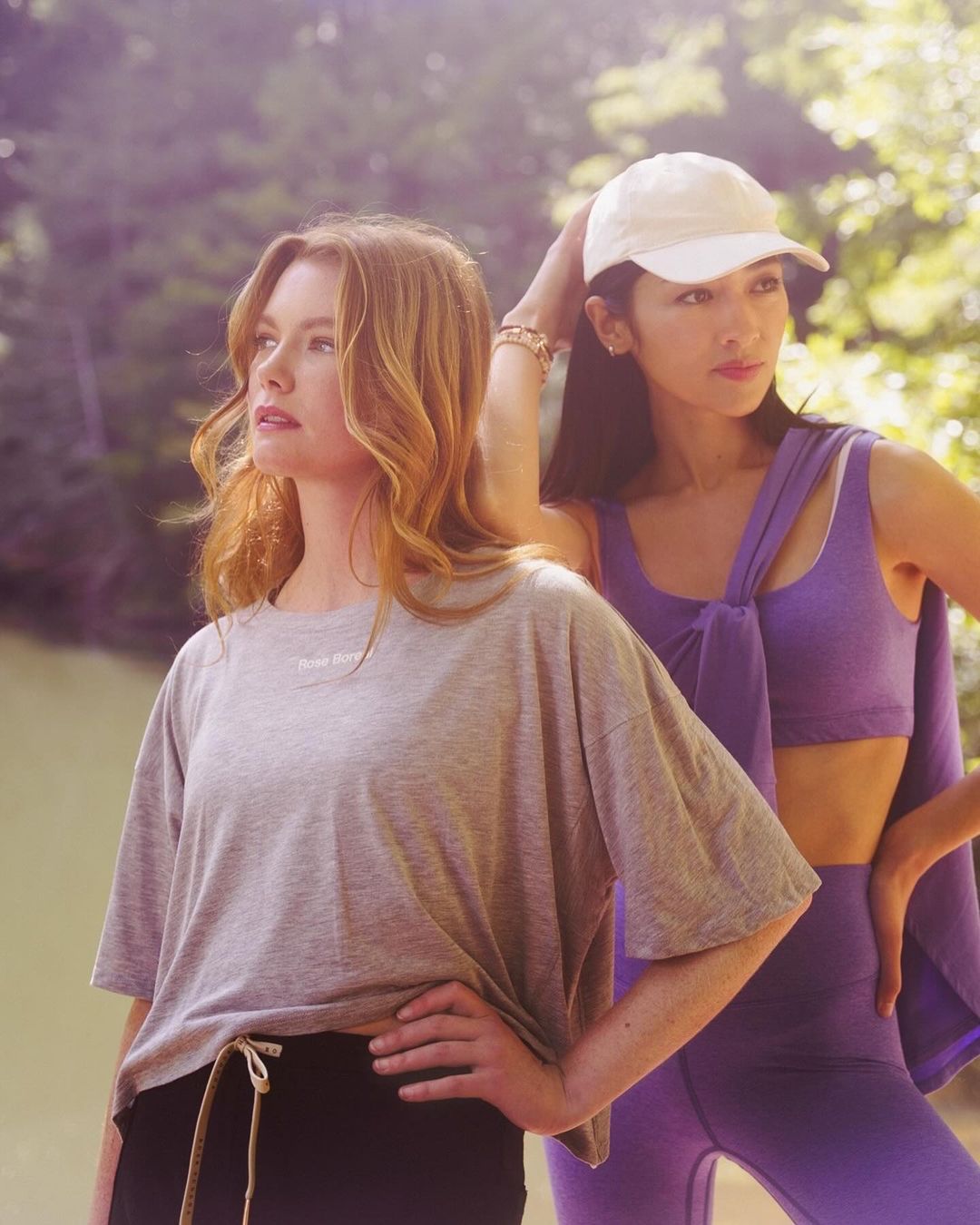 Two women pose in activewear outdoors. One in gray tee and black shorts, the other in purple leggings and a matching top. Natural lighting.
