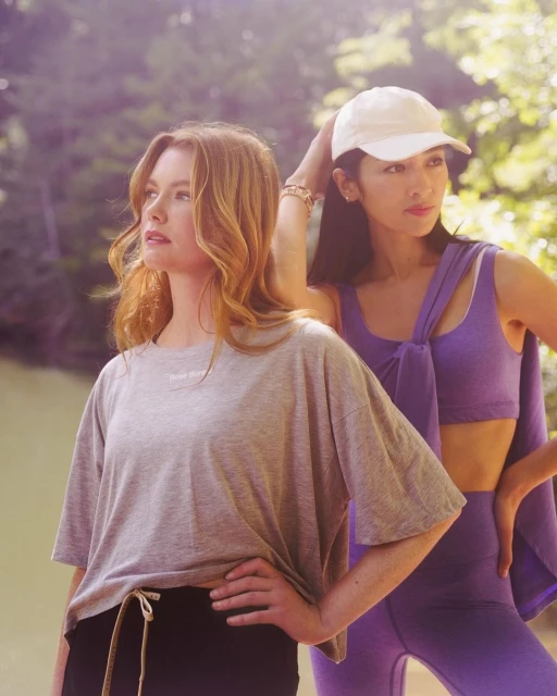 Two women pose in activewear outdoors. One in gray tee and black shorts, the other in purple leggings and a matching top. Natural lighting.