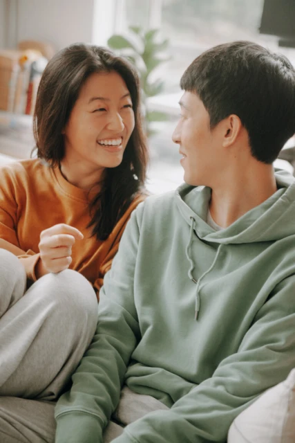 Young Asian couple smiling and looking at each other indoors. The woman is wearing an orange top, and the man is in a light green hoodie.