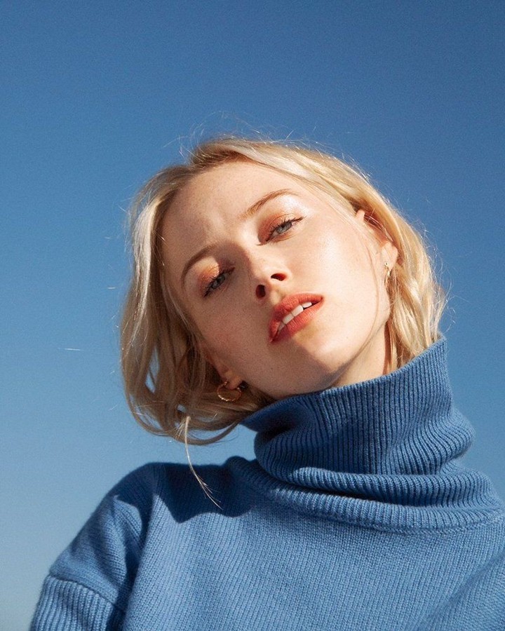 Close-up shot of a young, blonde adult woman wearing a blue turtleneck sweater against a clear blue sky background.