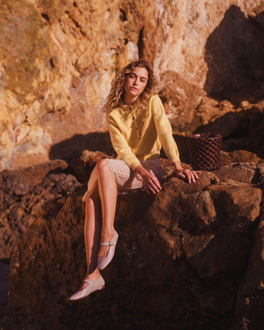 A young woman with curly hair sits on a rock, wearing a yellow cardigan and a polka dot skirt, accessorized with cream-colored shoes and a brown bag.