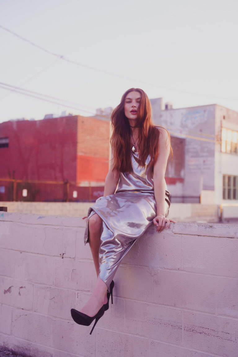 Young woman in silver dress and black high heels sits on a wall in an urban setting with buildings against a light blue sky.