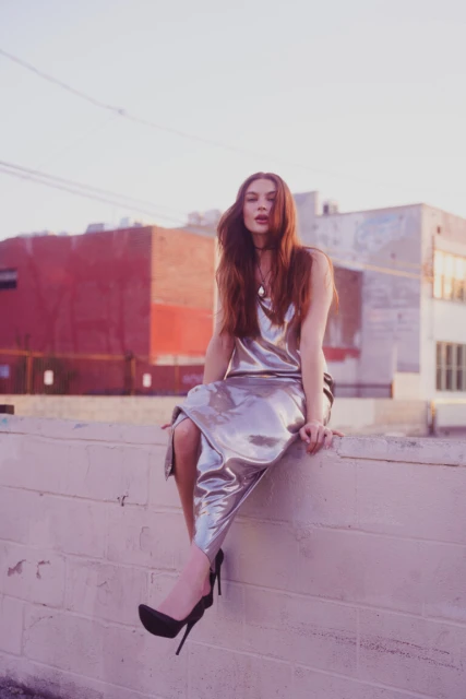 Young woman in silver dress and black high heels sits on a wall in an urban setting with buildings against a light blue sky.