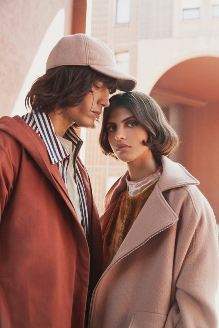 Close-up portrait of two young models wearing warm coats and baseball cap outdoors in front of terracotta architecture.