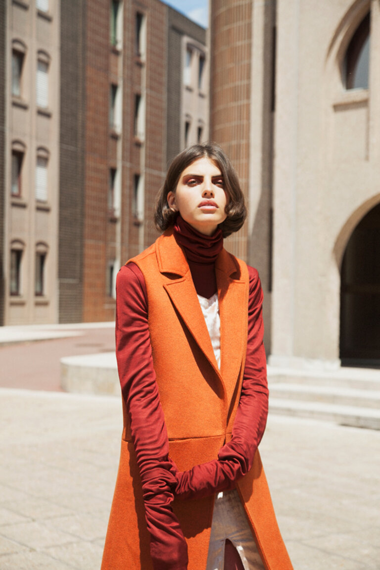 Fashionable adult woman poses in an outdoor urban setting, wearing a rust-colored vest, gloves and turtleneck, with modern buildings behind.