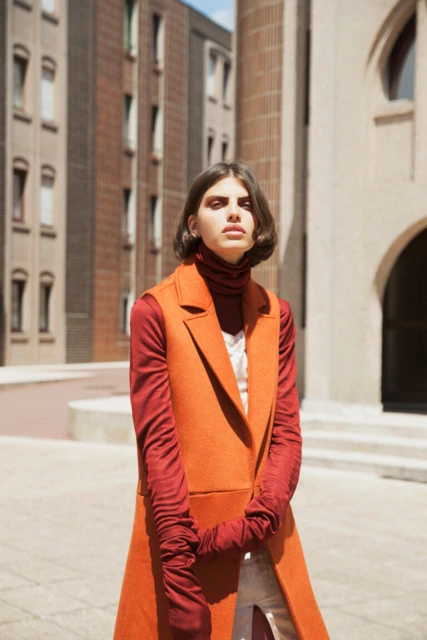 Fashionable adult woman poses in an outdoor urban setting, wearing a rust-colored vest, gloves and turtleneck, with modern buildings behind.