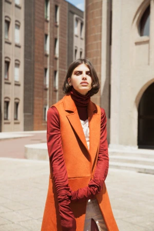 Fashionable adult woman poses in an outdoor urban setting, wearing a rust-colored vest, gloves and turtleneck, with modern buildings behind.