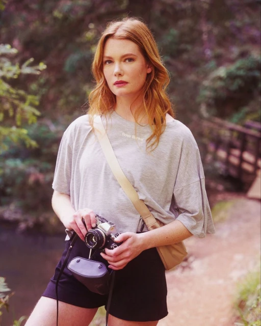 Red-haired woman in a forest setting, holding a vintage camera. She is wearing a grey t-shirt and black shorts.