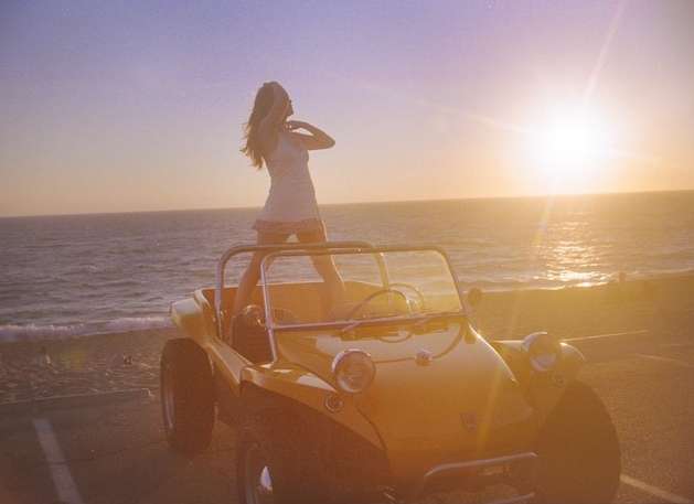 Woman standing on a yellow dune buggy at the beach with a sunset in the background, creating a serene and summery atmosphere.