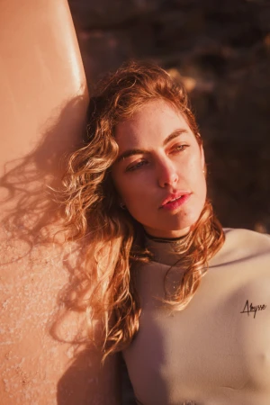 Close-up portrait of a woman with curly hair wearing a wetsuit leaning against a surfboard outdoors in warm sunlight.