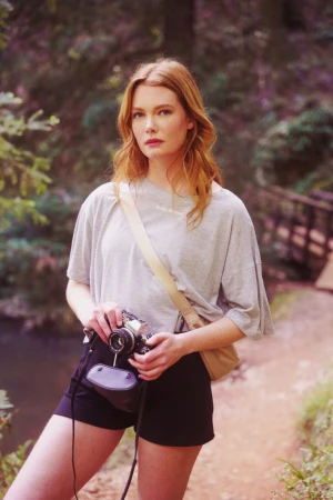 Red-haired woman holding a vintage camera wearing a grey t-shirt and black shorts on a wooded path by Nastasia Dusapin